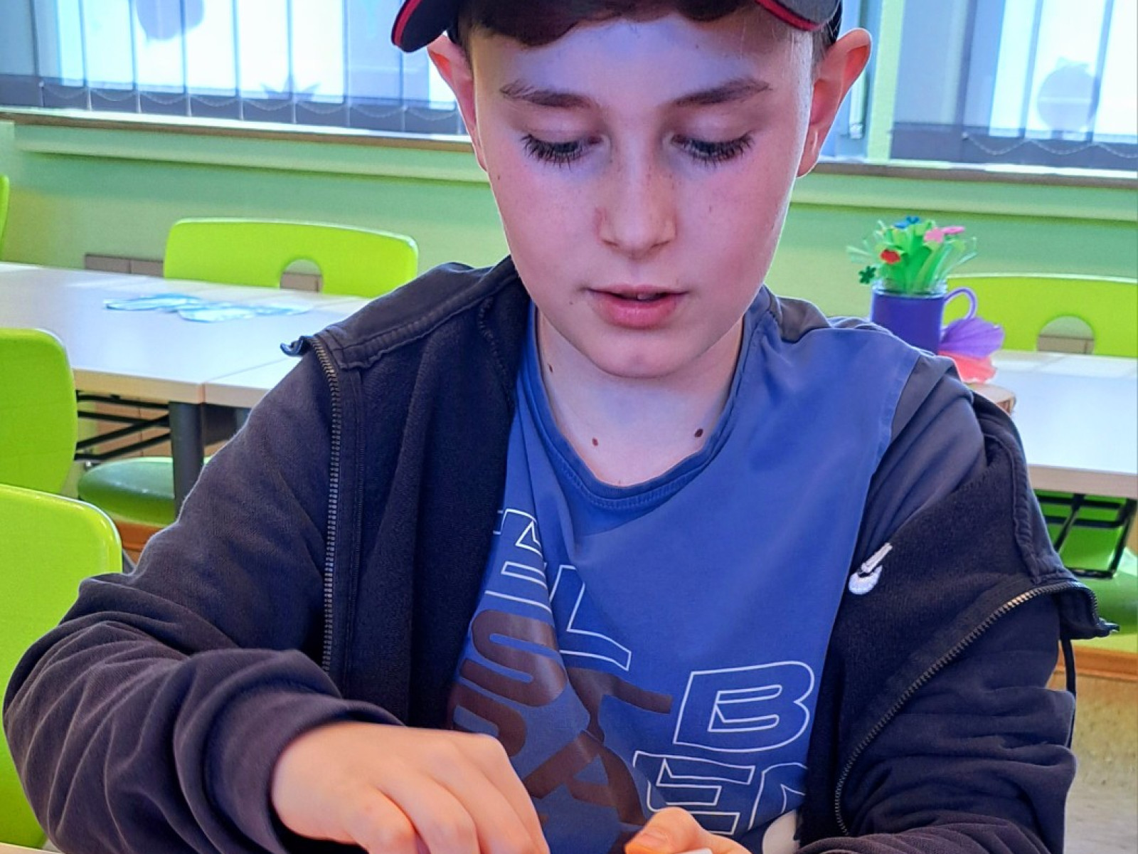 Junge mit Mütze isst aus einer Schale im Klassenzimmer, grüne Stühle im Hintergrund.