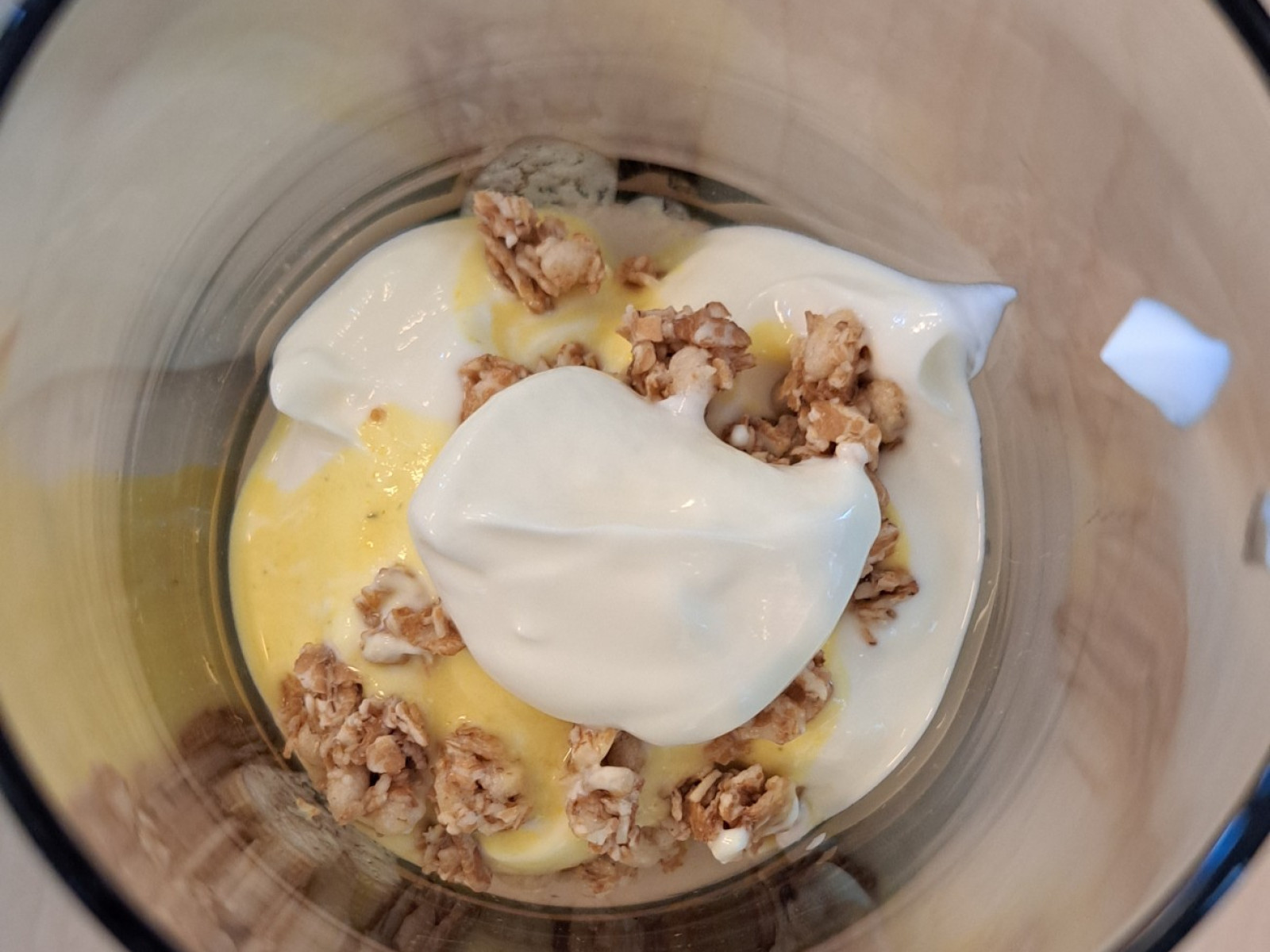 Glas-Schale mit Joghurt, gelbem Fruchtpüree und Müsli, von oben gesehen.