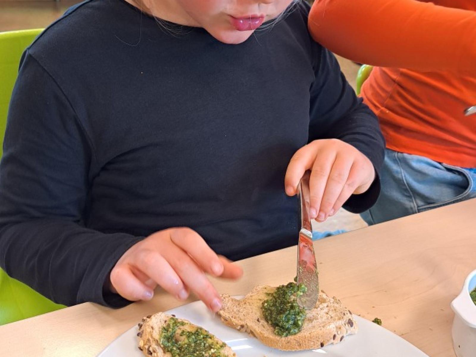 Kind bestreicht Vollkornbrotscheiben mit grünem Aufstrich, sitzt an einem Tisch.