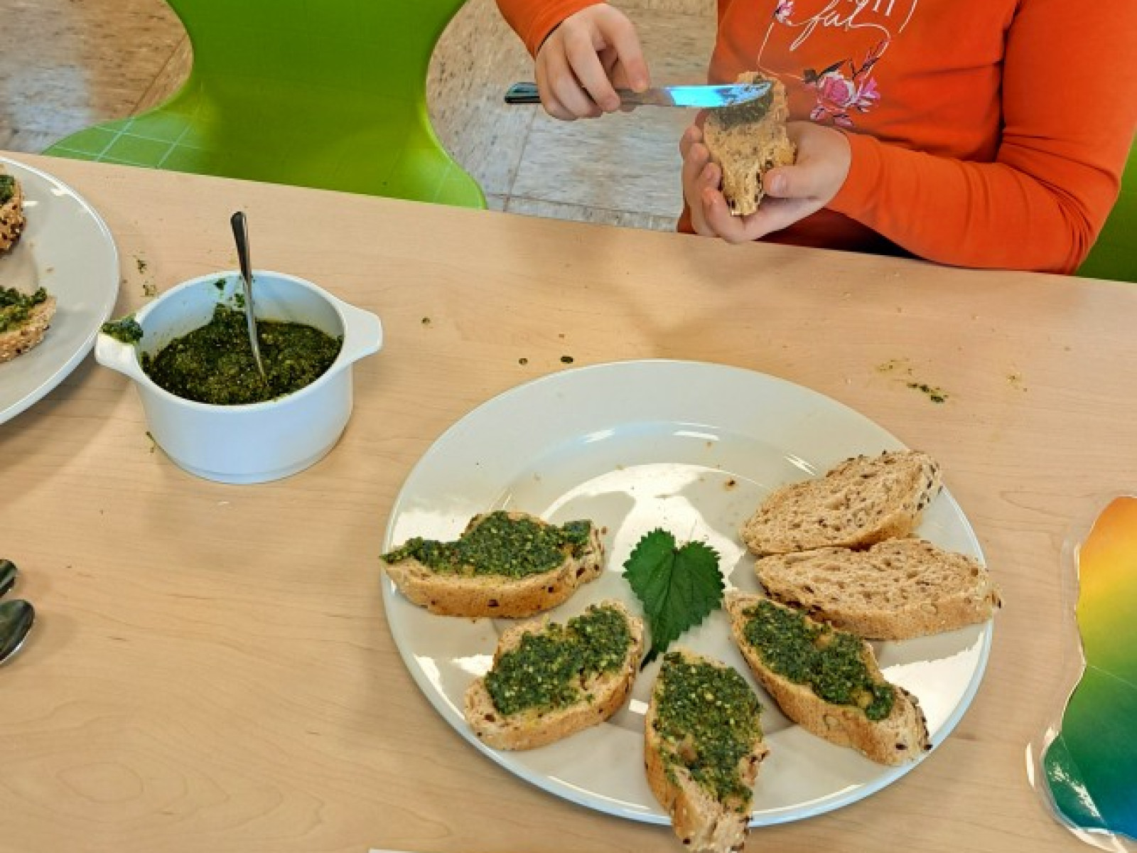 Kind streicht grüne Paste auf Brot, umgeben von weiteren Brotscheiben und einem Rezeptblatt.