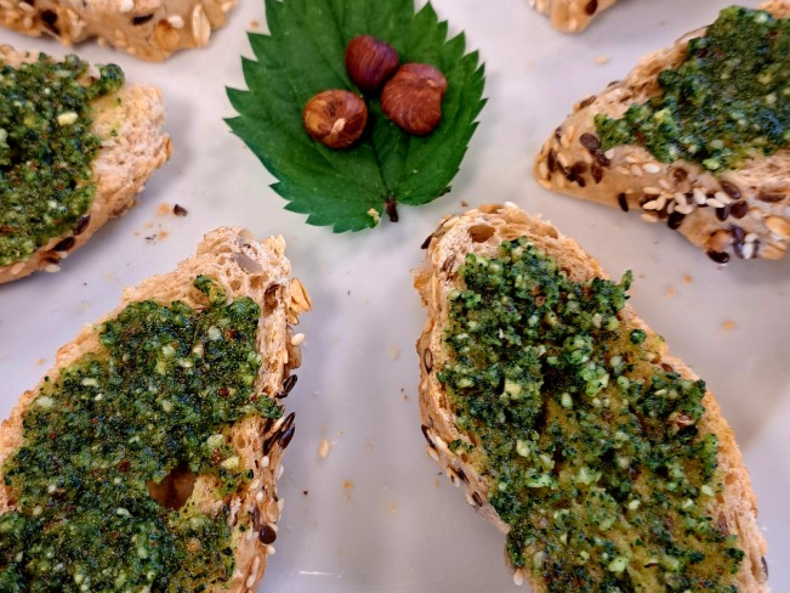Ein weißer Teller mit Scheiben Vollkornbrot, bestrichen mit grünem Kräuterpesto. In der Mitte des Tellers liegt ein Blatt mit zwei Haselnüssen.