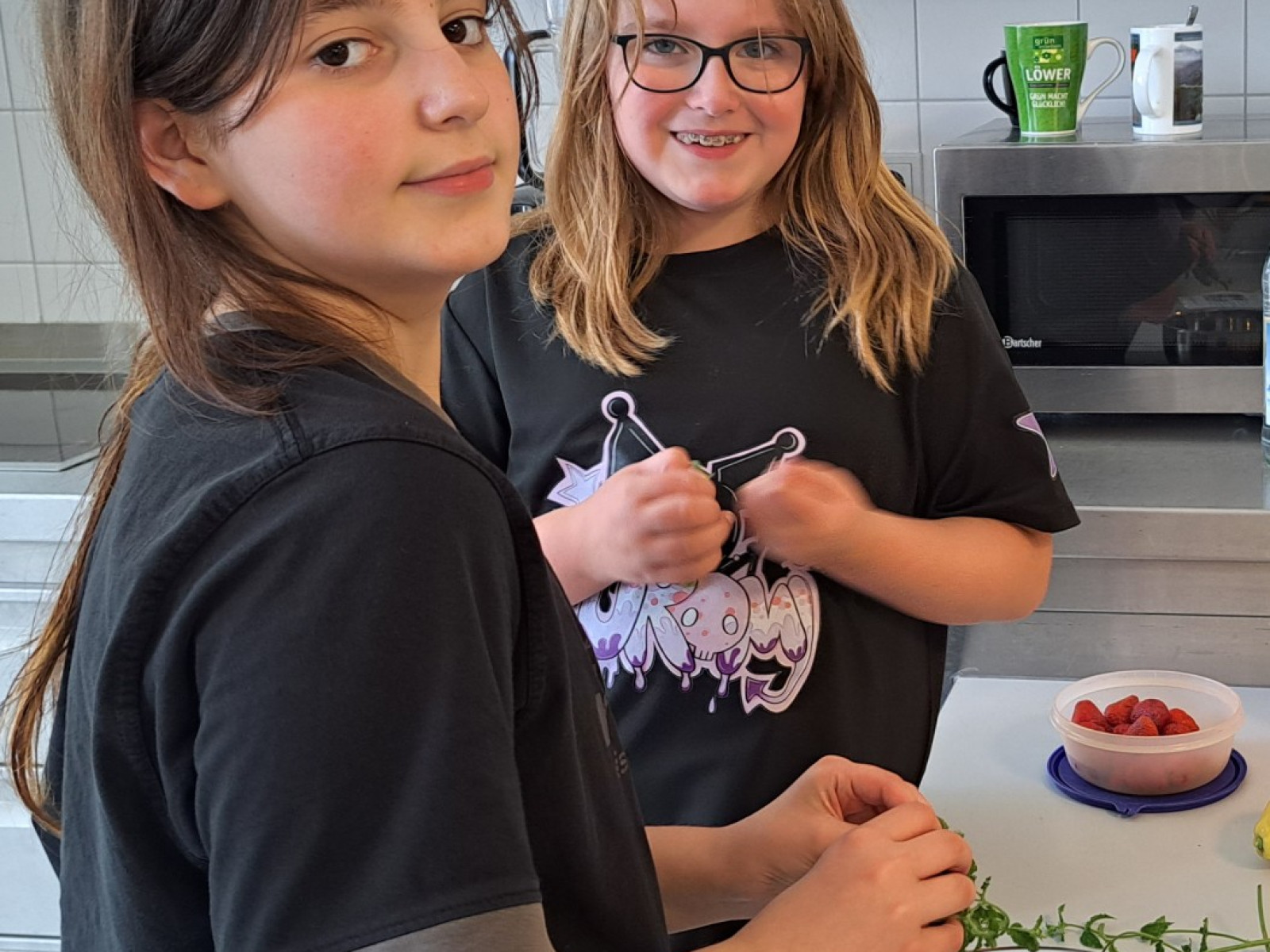 Zwei Kinder in einer Küche, die an einem Tisch arbeiten, frische Zutaten und Küchengeräte im Hintergrund.