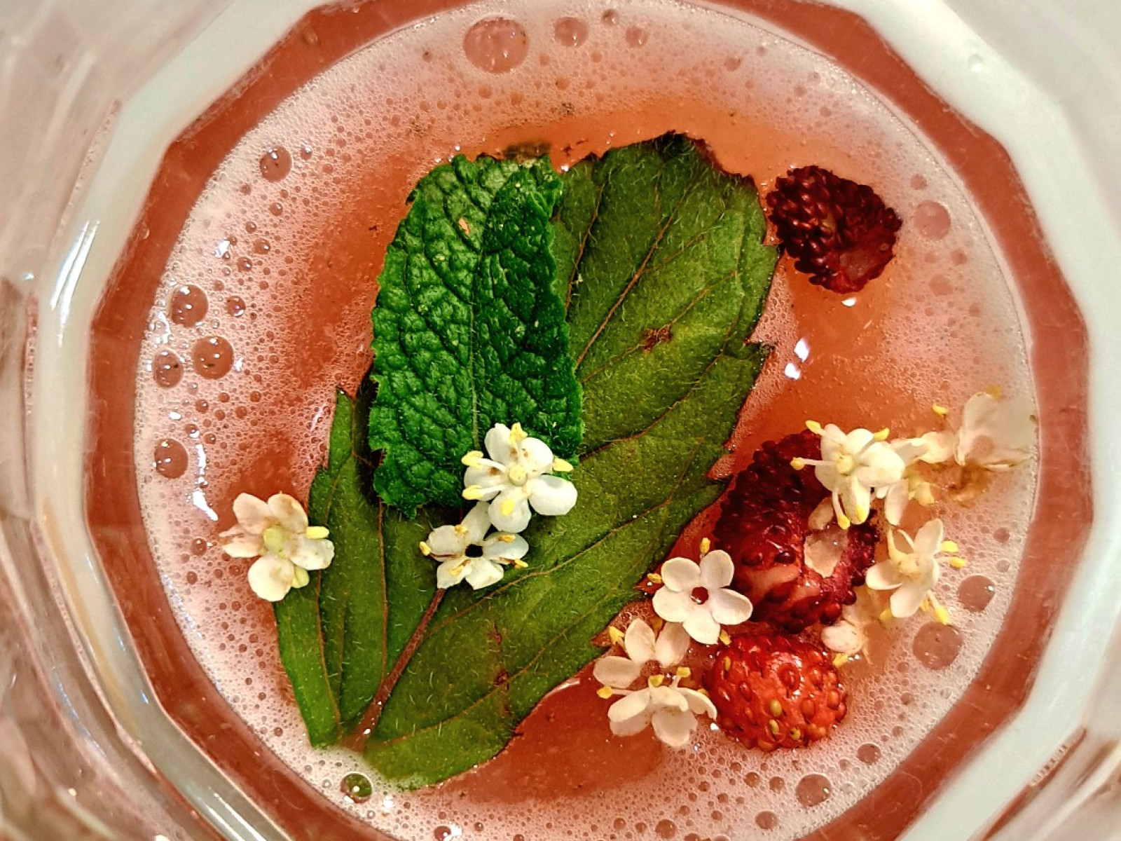Ein Glas mit rosafarbenem Getränk, garniert mit Minzblättern, Beeren und weißen Blüten.
