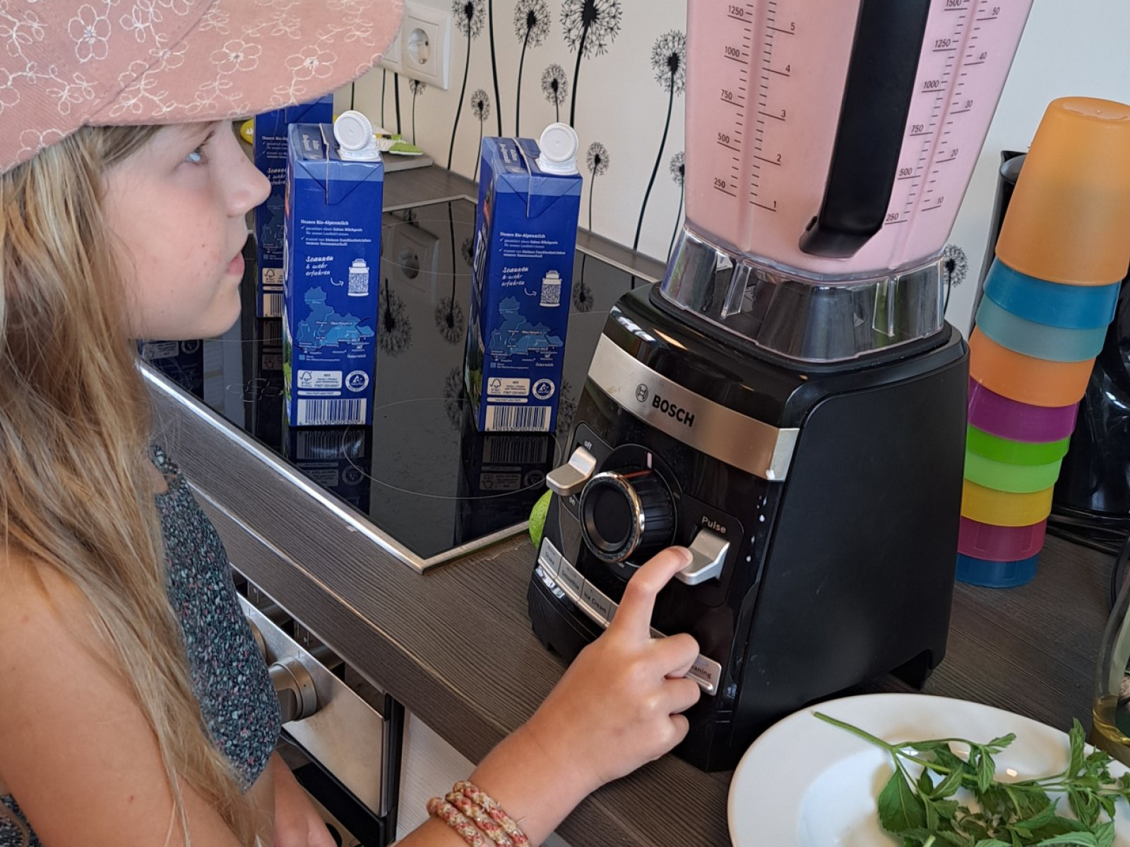 Ein Mädchen mit rosa Hut bedient einen Mixer mit rosa Inhalt. Daneben stehen Milchpackungen und ein Teller mit grünen Blättern.
