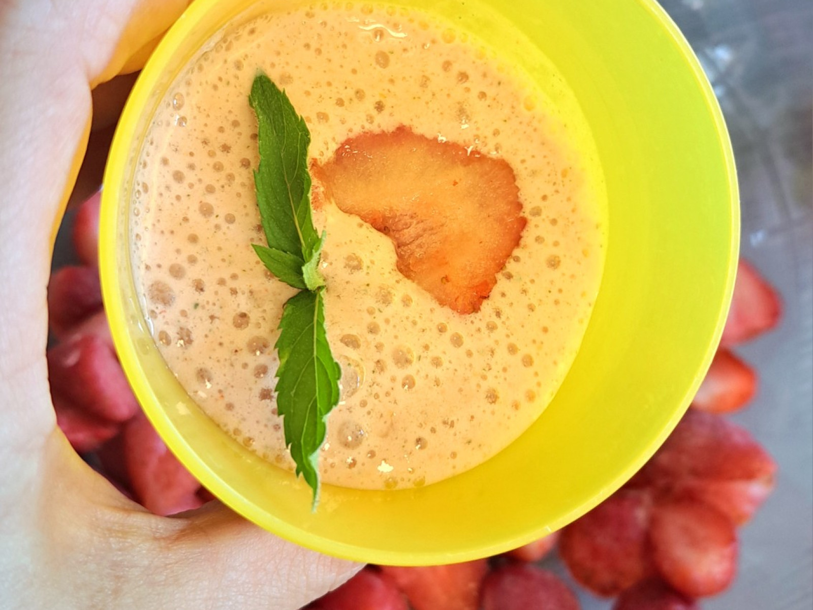 Gelber Becher mit Erdbeer-Bananen-Smoothie, garniert mit Erdbeere und Minze, vor geschnittenen Erdbeeren.