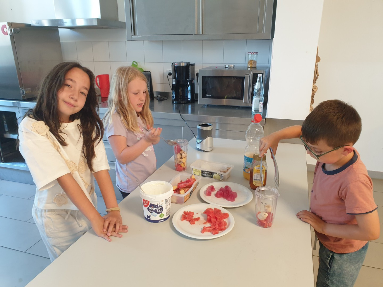 Drei Kinder stehen in einer Küche und bereiten Snacks mit Obst und Joghurt zu.