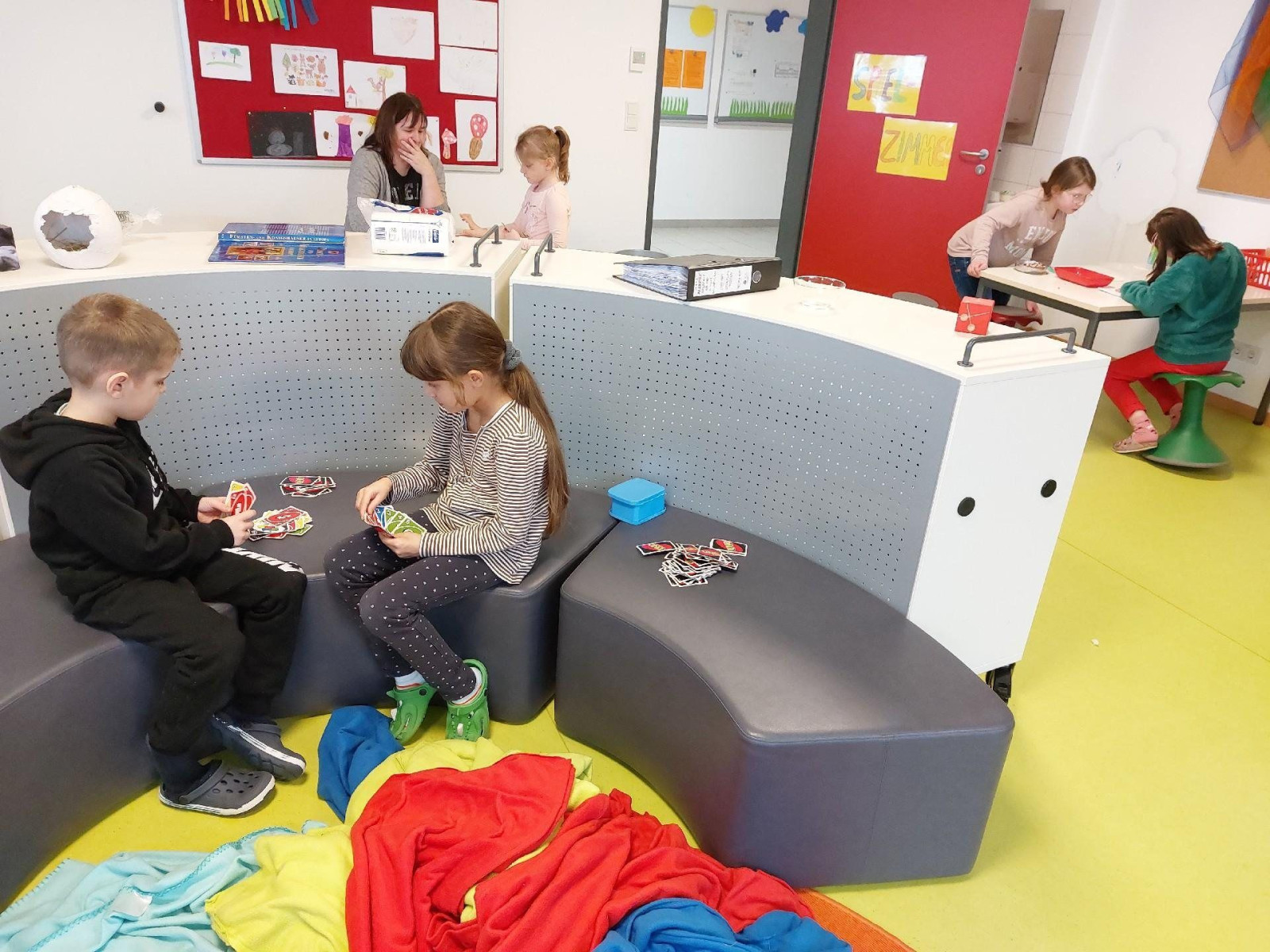 Kinder spielen in einem Klassenzimmer, sitzen auf Sofas und arbeiten an Tischen.