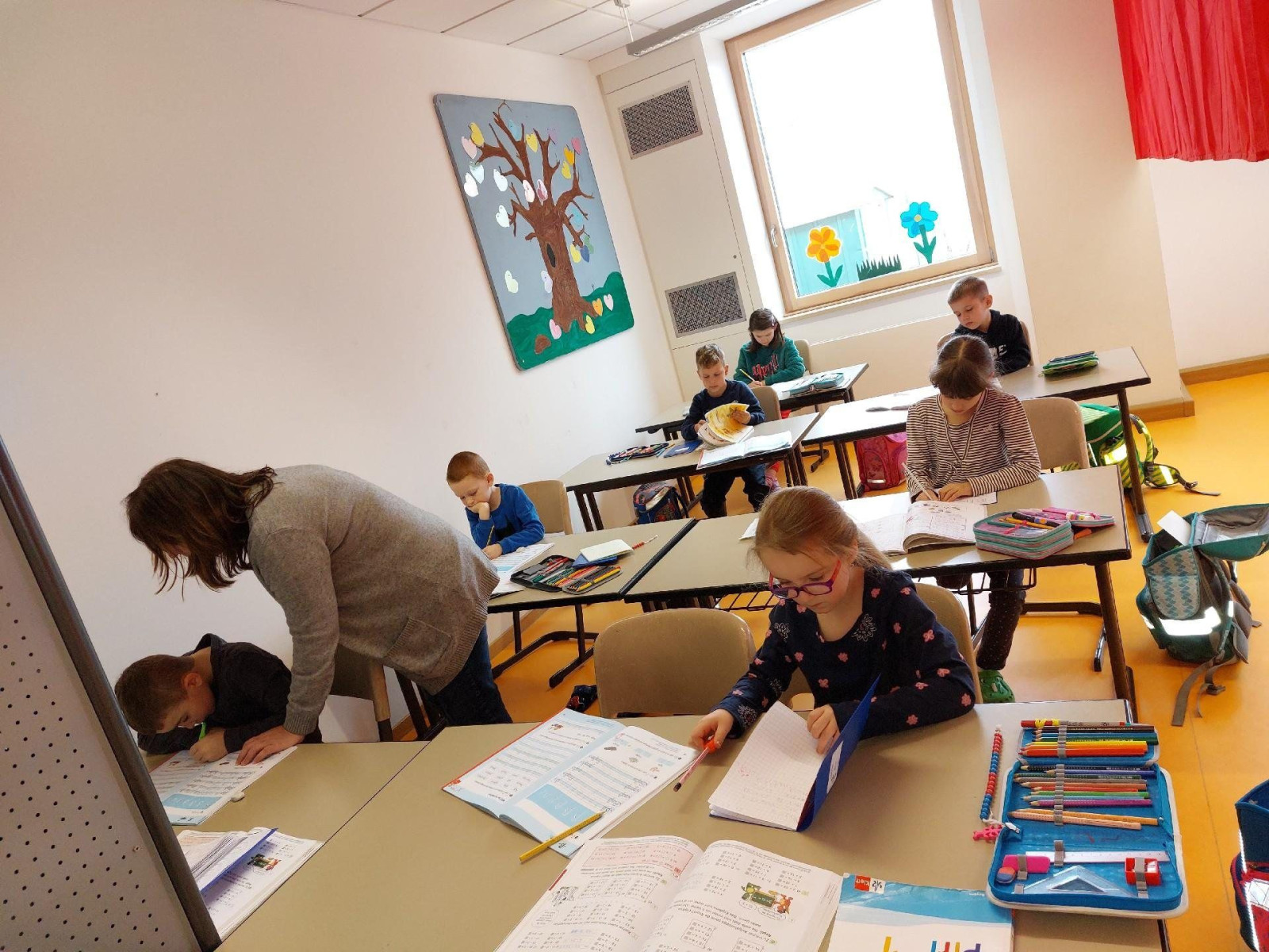 Lehrerin hilft Schülern in einem Grundschulklassenzimmer mit einem Baumplakat an der Wand.