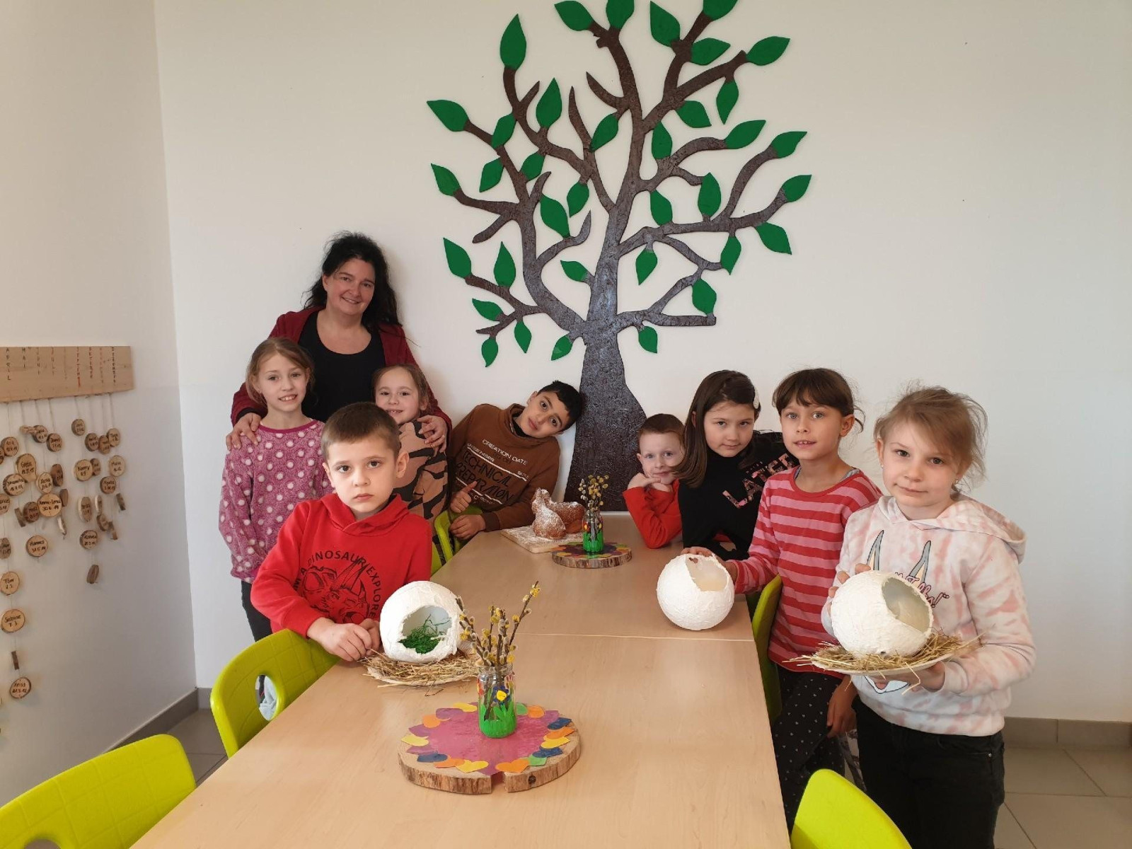 Kindergruppe um einen Tisch mit Bastelarbeiten, Wandbaum im Hintergrund.