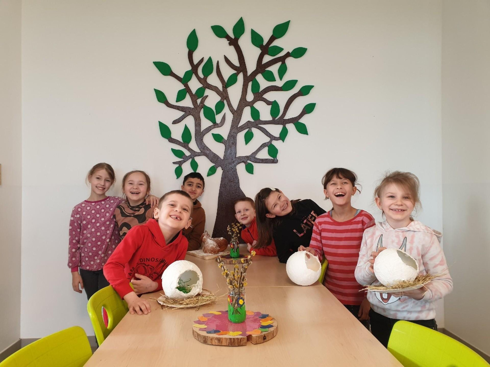Gruppe von Kindern lächelt vor einer baumförmigen Wanddekoration, an einem Tisch sitzend.
