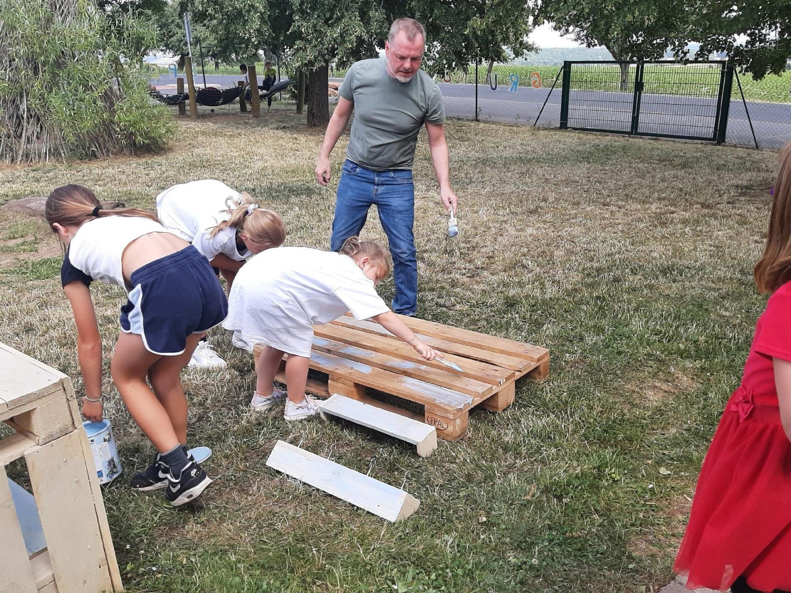 Kinder und ein Mann bemalen eine Holzpalette im Freien mit weißer Farbe.