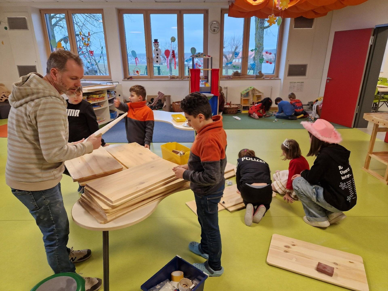 Kinder und ein Erwachsener arbeiten in einem Klassenzimmer mit Holzplatten. Der Raum ist farbenfroh dekoriert.