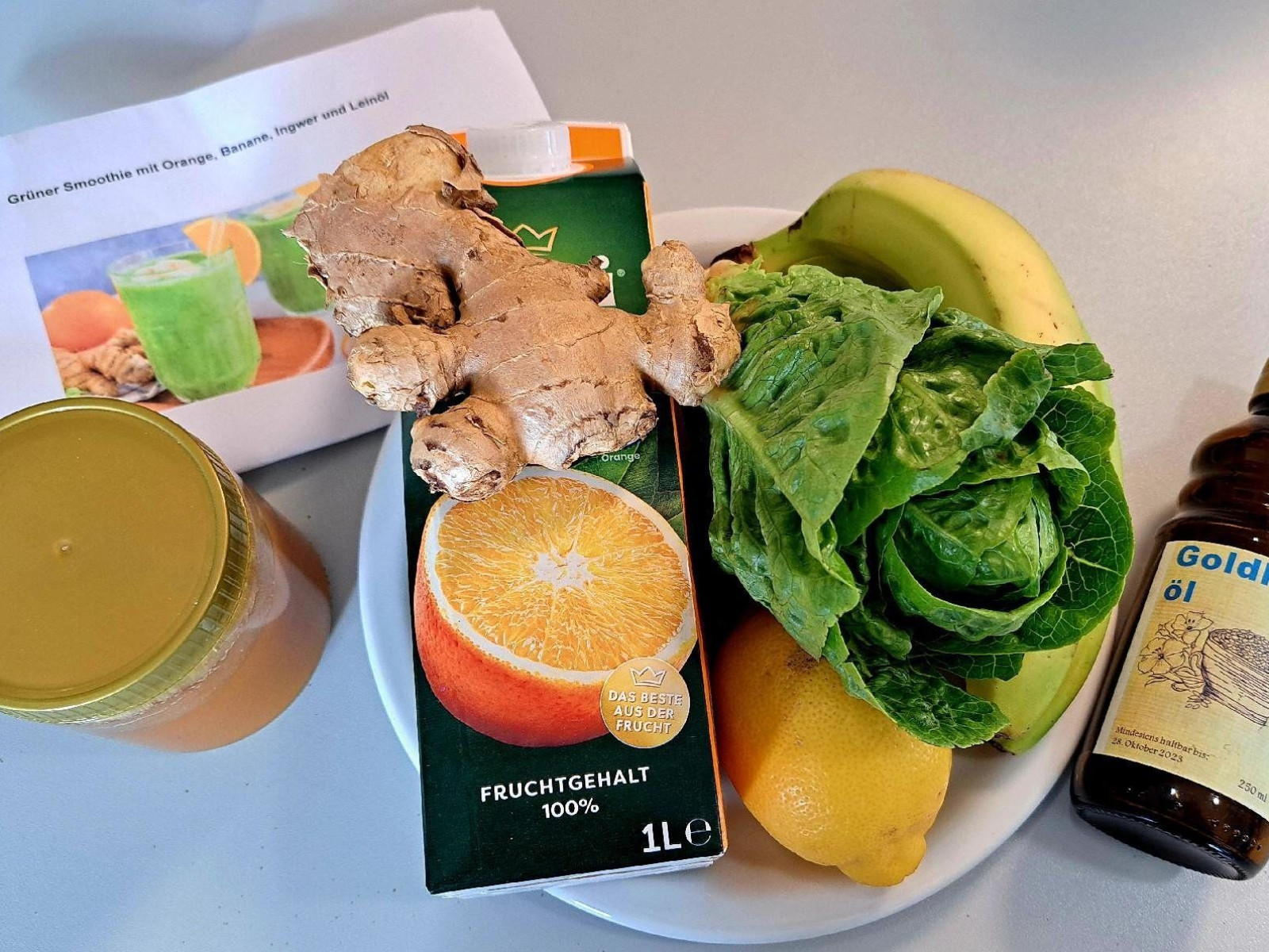 Ein Glas mit Saft, ein Rezeptblatt, ein Teller mit Orange, Ingwer, Banane, Zitronen, grüner Salat und eine Flasche Leinöl.