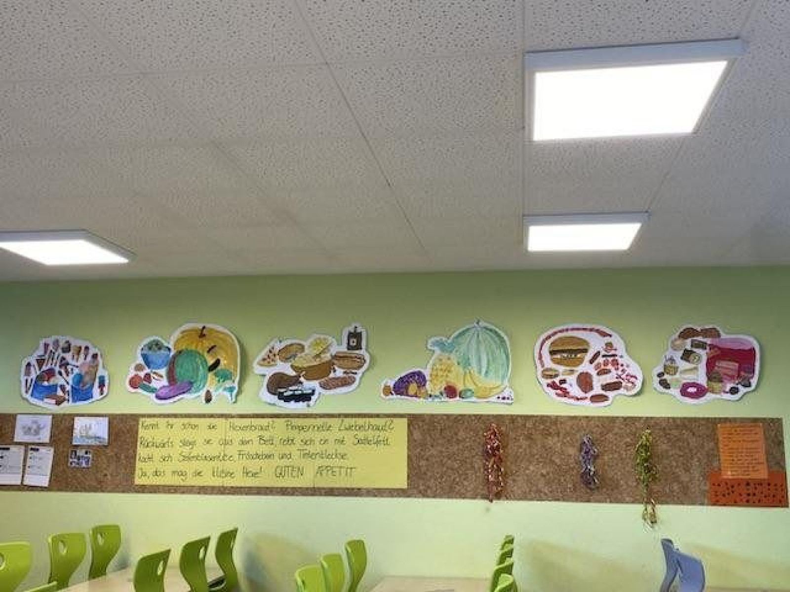 Klassenzimmerwand mit bunten Bildern von Lebensmitteln und grünen Stühlen davor.