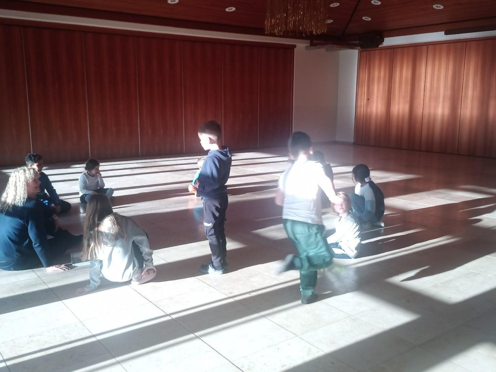 Kindergruppe sitzt und steht in einem Raum mit Holzverkleidung, Sonnenlicht fällt hinein.