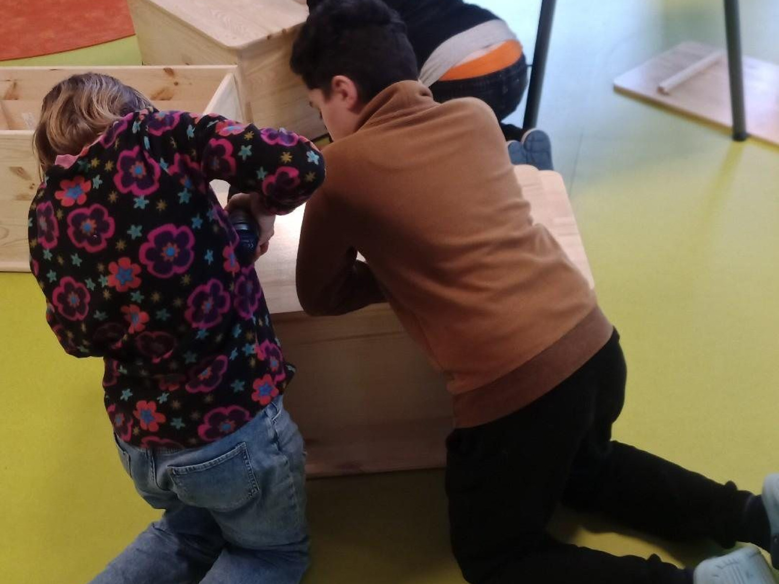 Kinder in einem Klassenzimmer knien auf dem Boden und bauen Holzkästen. Andere Kinder sitzen am Tisch.