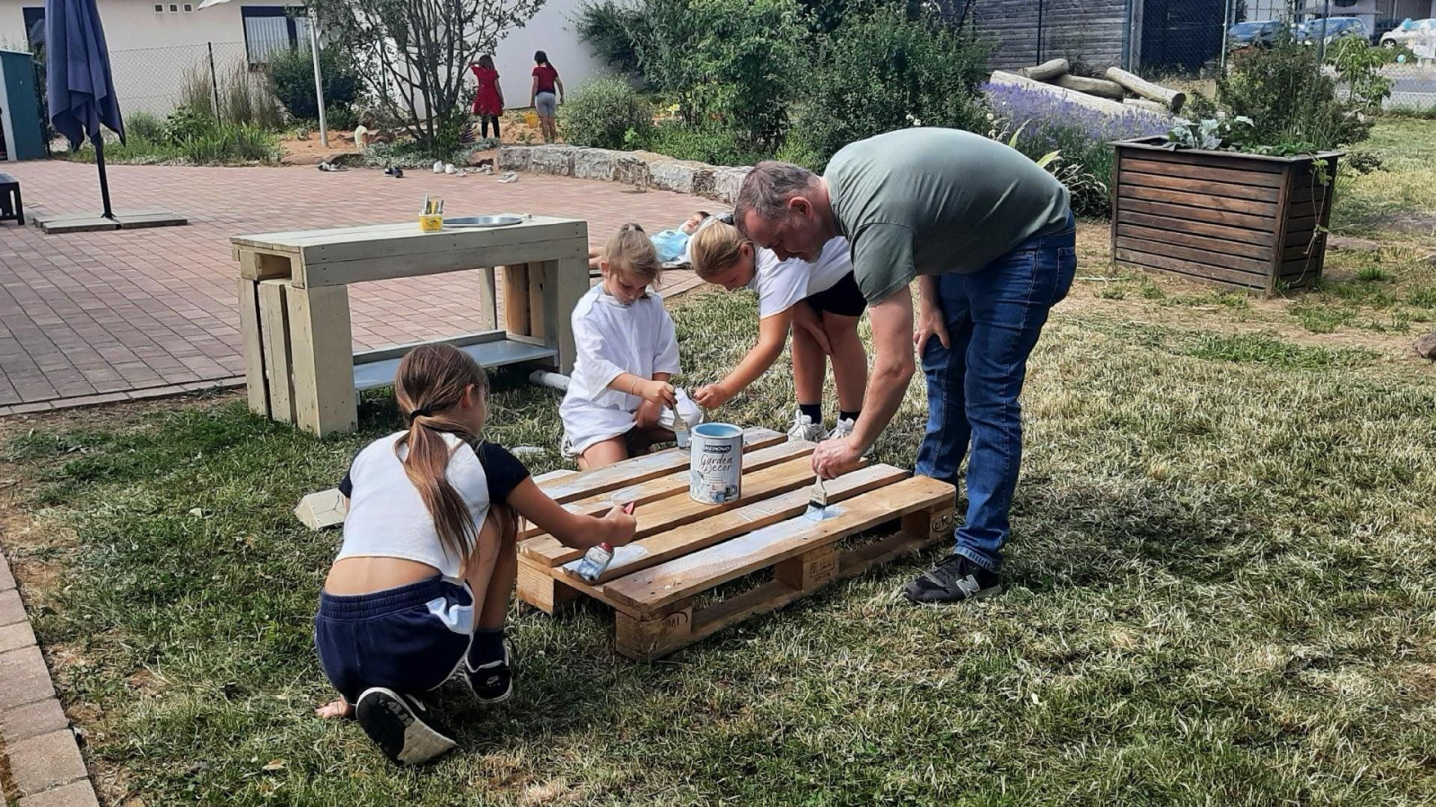 Eine Gruppe von Menschen streicht eine Holzpalette im Freien auf einer Grünfläche.