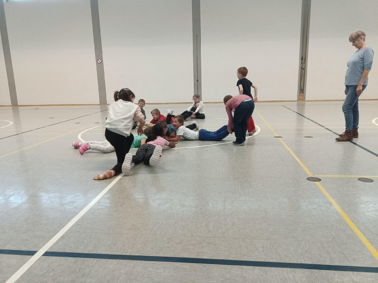 Kinder liegen und spielen auf dem Boden einer Turnhalle, während eine Frau zuschaut.
