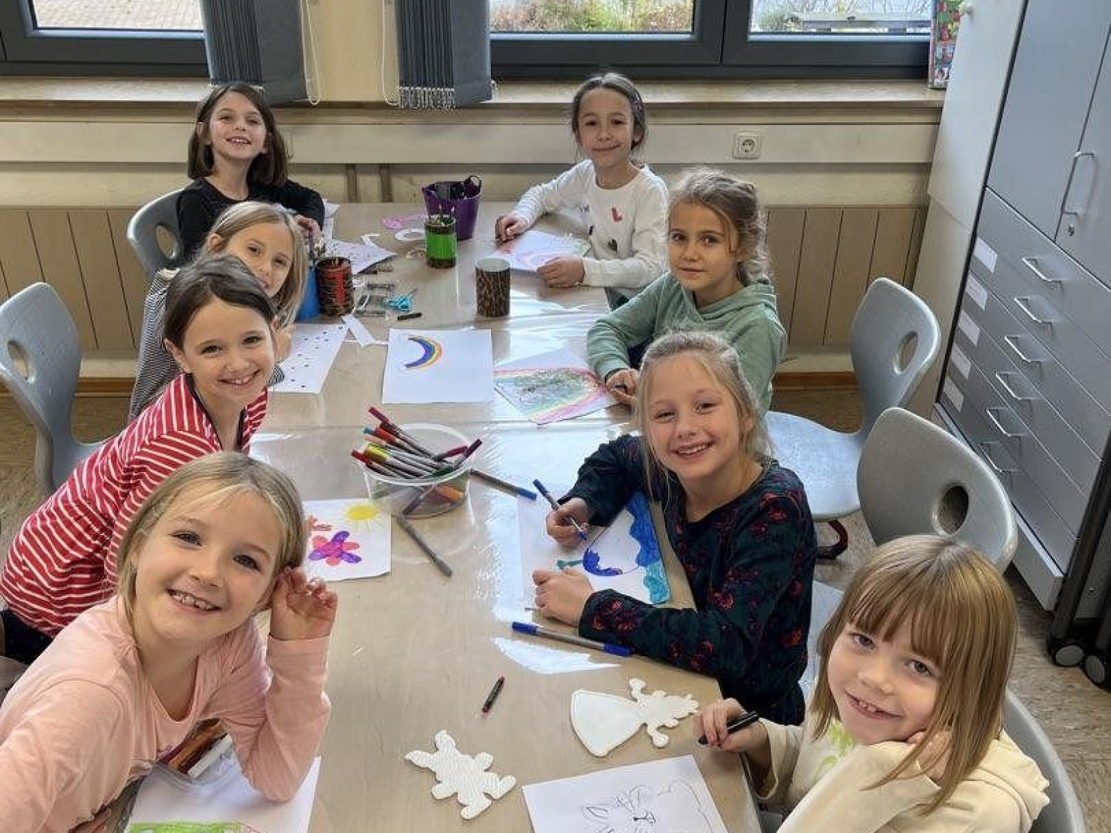 Kinder sitzen um einen Tisch und basteln im Klassenzimmer mit Papier und Stiften.