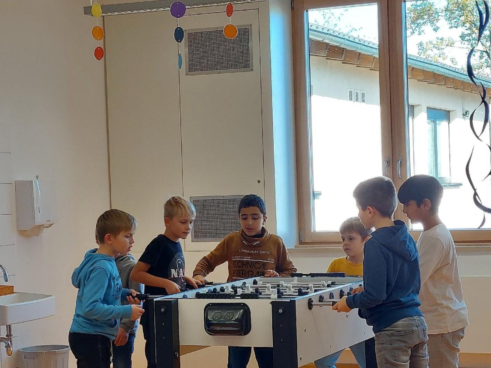 Sechs Kinder um einen Tischfußballtisch in einem Klassenraum mit bunter Dekoration.
