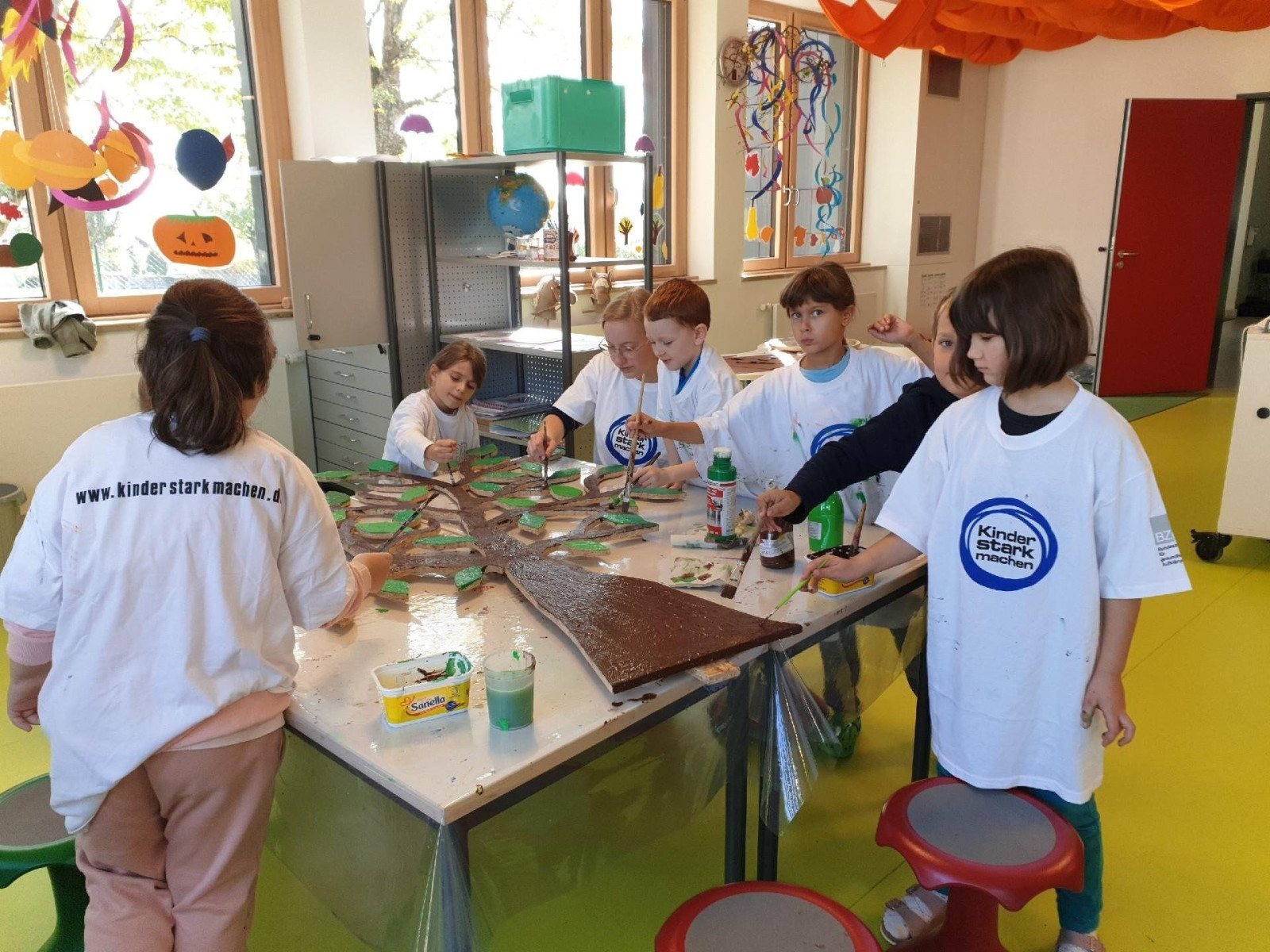 Kinder im Klassenzimmer basteln mit Materialien auf einem Tisch, T-Shirts der Startnetzwerk trägt.