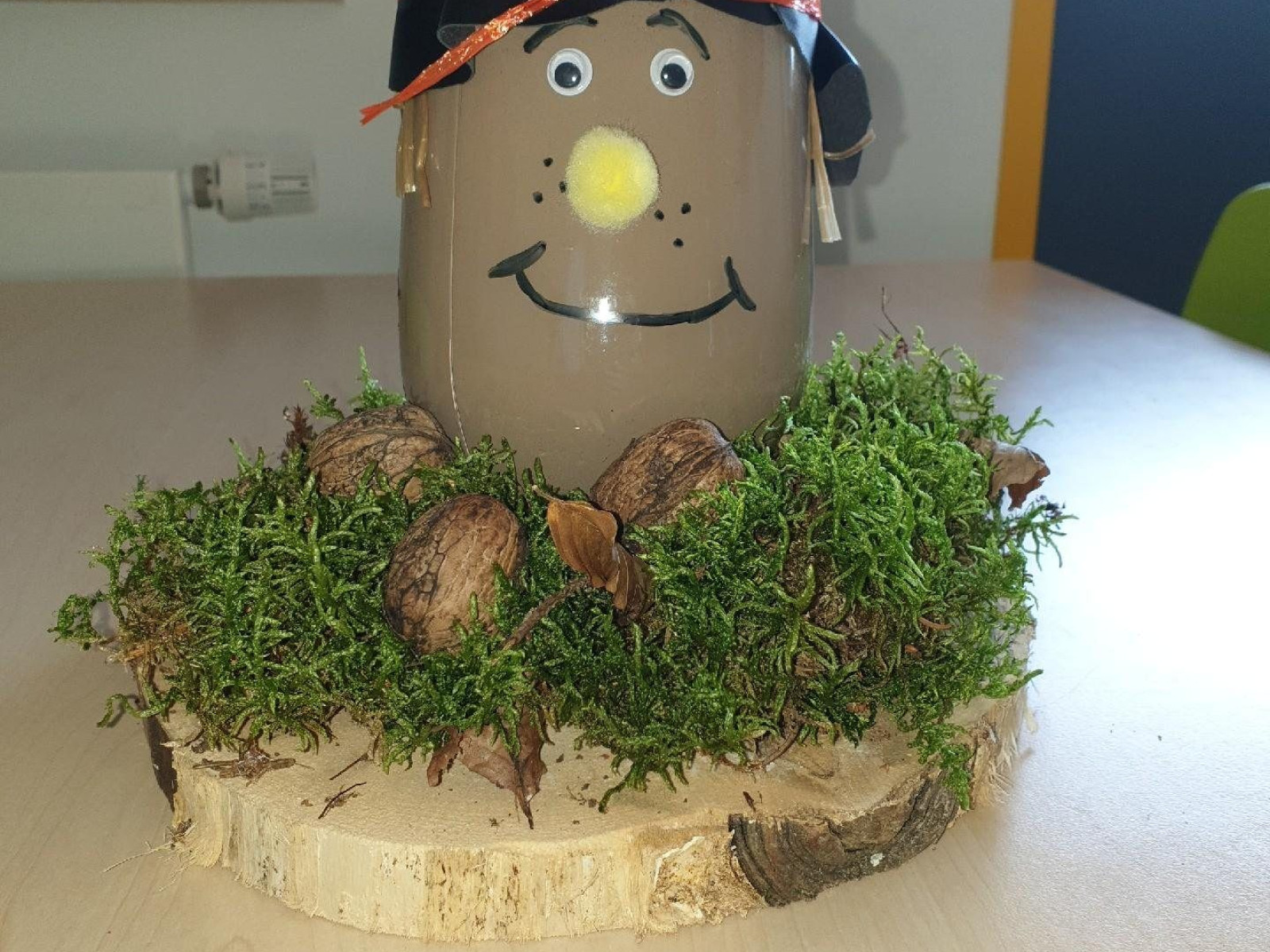 Ein Glas mit gemaltem Gesicht, Hut und oranger Schnur, umgeben von Moos und Nüssen auf Holz.